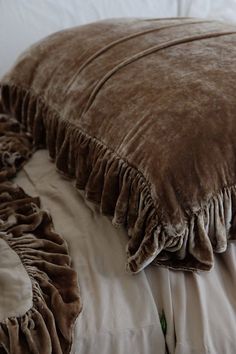 a brown blanket is laying on top of a white bed with ruffles and pillows