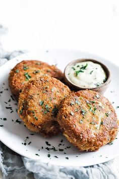 three crab cakes on a white plate with a small bowl of sour cream in the background