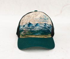 a green hat with a mountain scene on the front, and mountains in the back