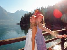 two girls are standing on a bridge near the water and trees, with mountains in the background