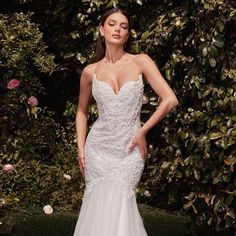 a woman in a white wedding dress standing next to some bushes and flowers with her hands on her hips