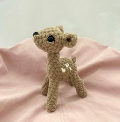 a small crocheted deer sitting on top of a pink sheet with black eyes