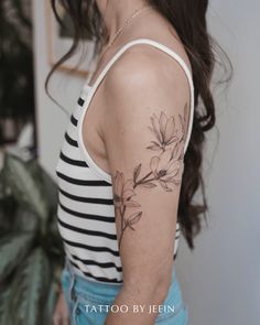 a woman with a flower tattoo on her left upper arm and shoulder, standing in front of a plant