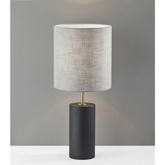 a black table lamp with a white shade on the top and a gold base, in front of a gray wall