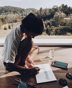 a man sitting on the floor writing in front of a window with an open notebook