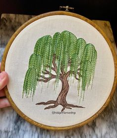 someone is holding up a small embroidery project with a tree in the center and green leaves on it