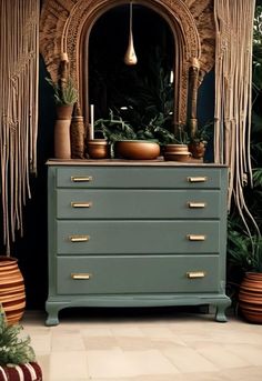 a green dresser with gold handles and fringes on it's sides, next to potted plants