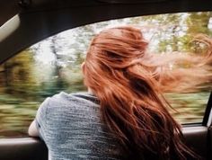 a woman with long red hair sitting in a car looking out the window at trees