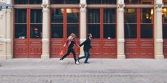 two people are walking down the sidewalk in front of a building with red doors and windows