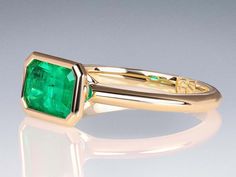 an emerald ring is shown on a reflective surface, with the center stone set in yellow gold