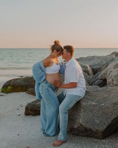 a pregnant couple sitting on rocks at the beach