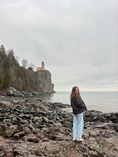 a woman standing on top of a rocky beach next to the ocean with a lighthouse in the background