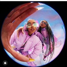 two women in pink jackets and sunglasses are seen through a circular lens with the reflection of one woman's hand