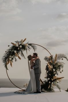 a bride and groom standing under an arch with flowers on the beach in front of the ocean