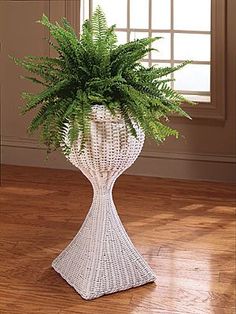 a white wicker vase with a green plant in it on a wooden floor next to a window