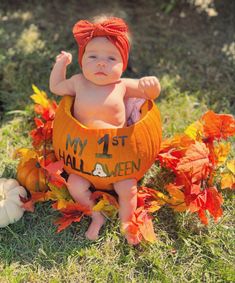 a baby in a pumpkin costume sitting on the ground with leaves around him and his hand up