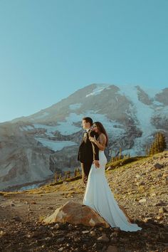 a bride and groom standing on top of a rocky hill with mountains in the background