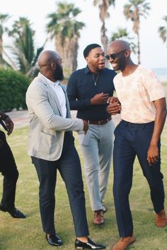 three men standing next to each other on top of a grass covered field with palm trees in the background