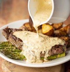 a person pouring sauce onto a plate of food with asparagus and potatoes on the side