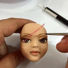 a person is holding a doll head with a needle in it's mouth and another hand holds a pen