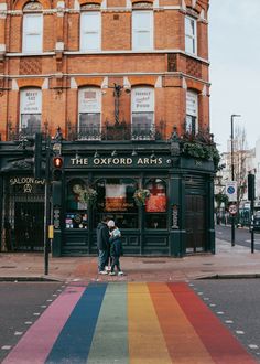 two people standing in front of a building with a rainbow painted crosswalk on the sidewalk