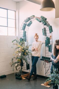 two women standing next to each other in front of a wall with plants on it