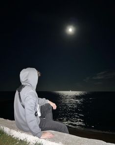 a man sitting on the edge of a cliff looking at the ocean under a full moon