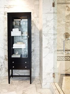 a bathroom with marble walls and flooring has a glass cabinet in the corner that holds towels