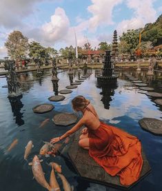 a woman in an orange dress kneeling down to catch fish from the water with her hands