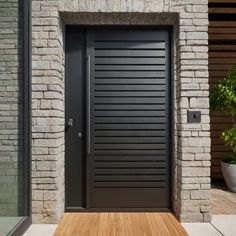 a black door is in front of a brick wall and wooden flooring with potted plants on either side