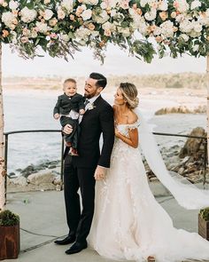 a bride and groom holding their son under an arch with flowers on the beach in front of them