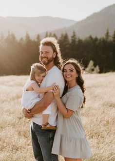 a man and woman holding a baby in a field with mountains in the background at sunset