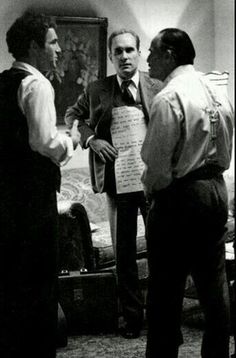 three men standing in a living room talking to each other and one man holding a piece of paper