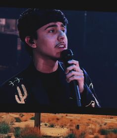 a young man holding a microphone in front of a tv screen with desert scene behind him