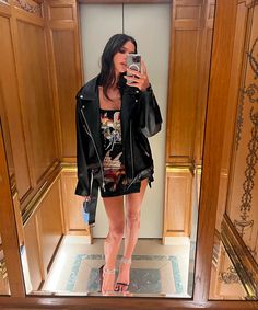 a woman taking a selfie in front of a mirror wearing high heels and a leather jacket