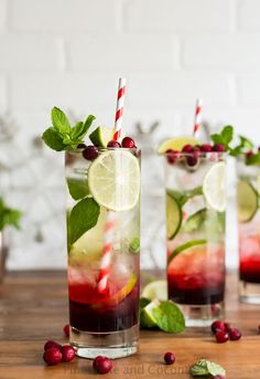 two glasses filled with drinks and garnished with limes, cranberries, and mint