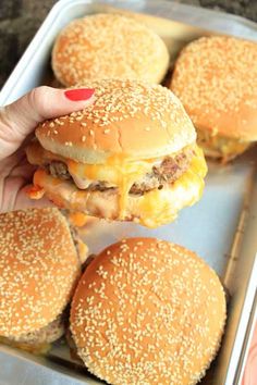 a person holding a hamburger in front of some other hamburgers on a baking sheet