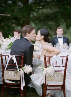 a bride and groom kissing in front of their guests at the dinner table with chairs