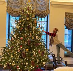 a woman standing next to a christmas tree in a room with large windows and curtains