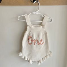 a white knitted baby romper with the word one on it hanging from a hook