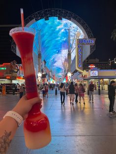 a person holding up a drink in front of a building with lights and people walking around