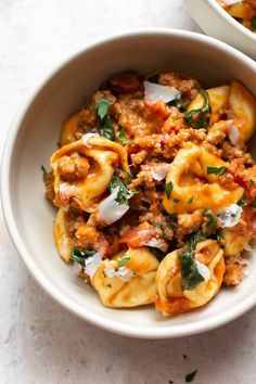 a white bowl filled with pasta and meat covered in sauce, parmesan cheese and herbs