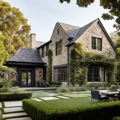 a large stone house surrounded by greenery and trees with an outdoor dining table in the foreground