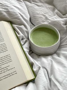 an open book sitting on top of a bed next to a bowl of green liquid