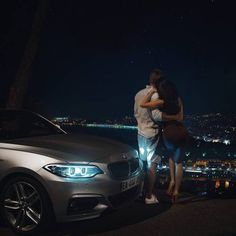 a man and woman standing next to a silver car at night with city lights in the background