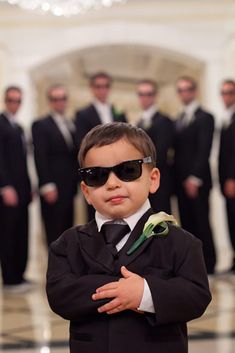 a young boy wearing sunglasses and standing in front of a group of other men with their arms crossed