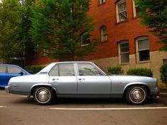 two cars are parked on the street in front of a brick building with trees and bushes