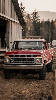 an old red pick up truck parked in front of a barn with a wooden building