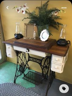 an antique sewing table with two vases and a plant on top, in front of a yellow wall