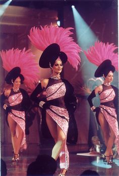 two women in pink dresses and black hats are dancing on stage with bright lights behind them